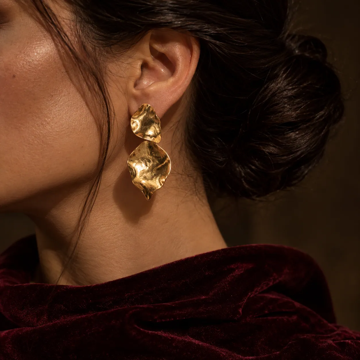 Orecchino d’oro martellato su un collo di velluto bordeaux. Persona inventata, immagine generata con AI.
